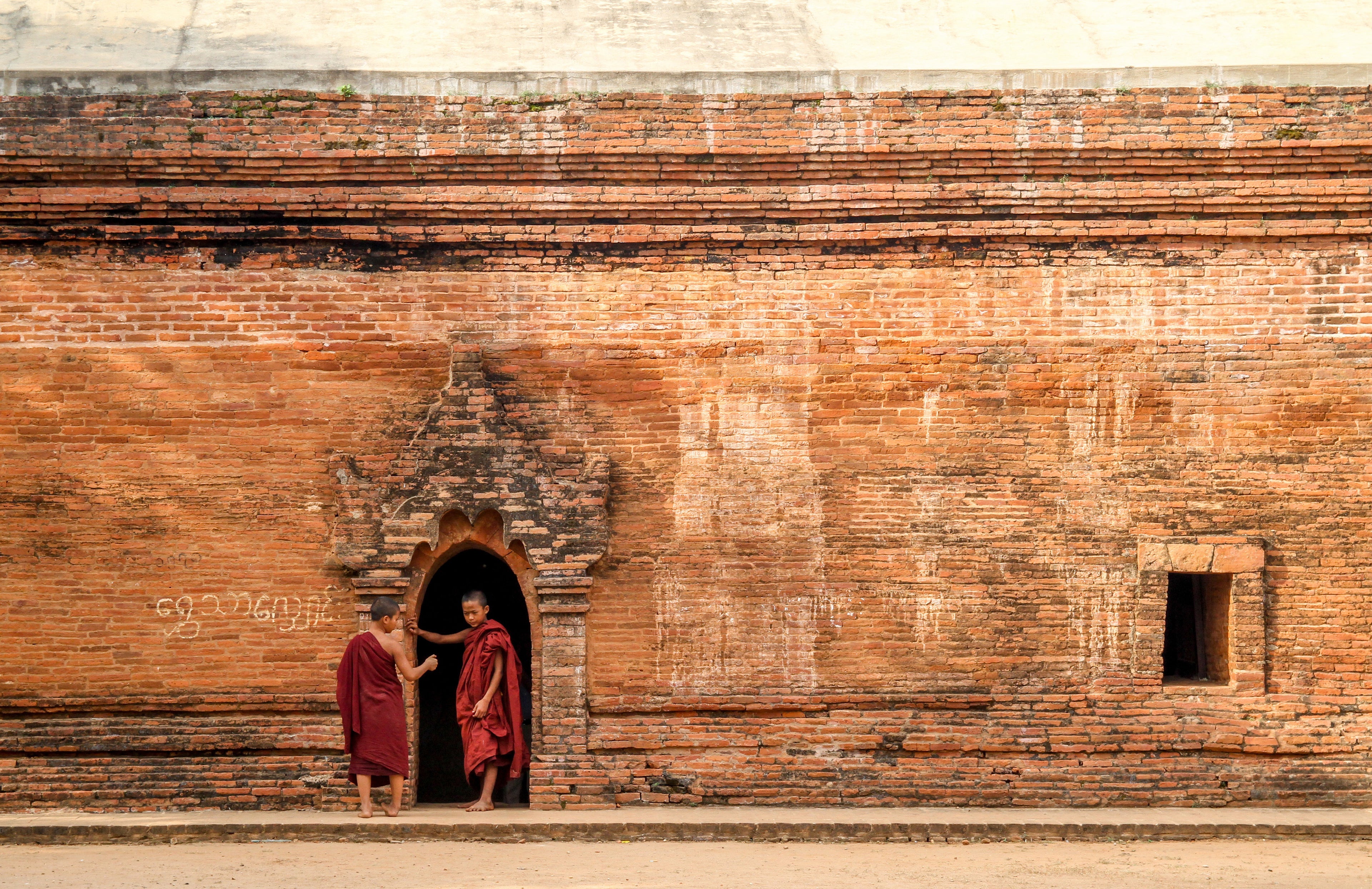 Two monks in red robes standing in front of a large brick wall with a doorway.